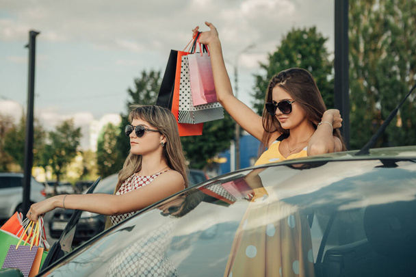 Портрет красивых симпатичных чирлидерш, идущих домой после шоппинга, стоящих рядом с машиной, радуясь новым вещам носить желтые точечные кроссовки платье обувь черная пятница концепция - Фото, изображение