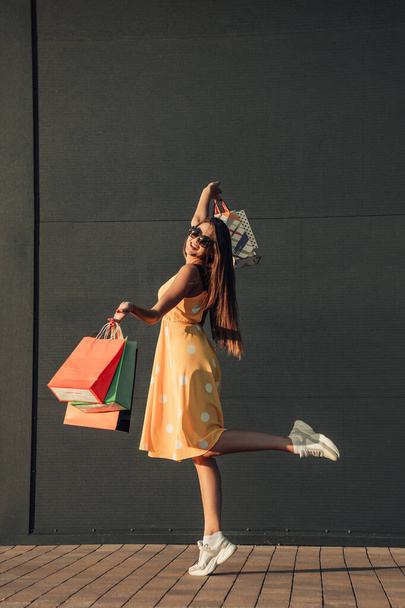Pionowa pełna długość ciała portret młodej uśmiechniętej dziewczyny ubranej w letnie ubrania stojącej na jednej nodze i korzystającej z zakupów torby z zakupami. - Zdjęcie, obraz