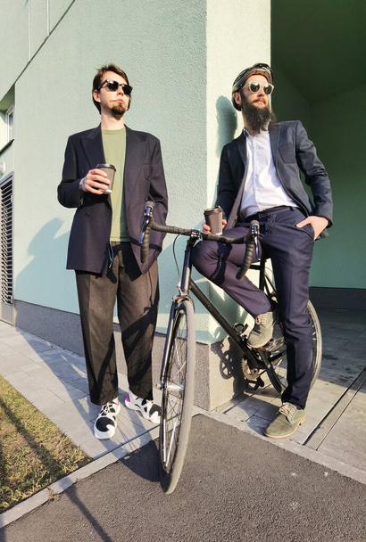 Two young men friends dressed casually spending time together at the city, drinking takeaway coffee next bike. - Fotó, kép