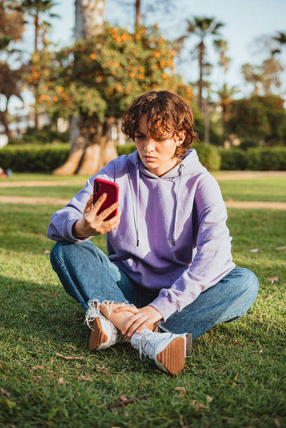 公園で携帯電話を見ている紫のパーカーを着た怒っているティーンエイジャー。彼女は草の中に座っている. - 写真・画像