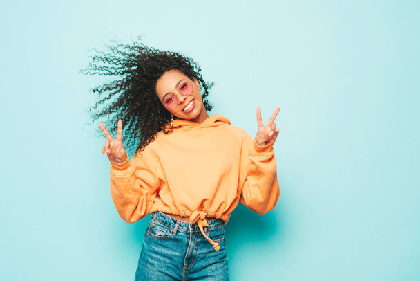 Beautiful black woman with afro curls hairstyle.Smiling model in orange hoodie and trendy jeans clothes. Sexy carefree female posing near blue wall in studio in sunglasses.Shows peace sign - Foto, afbeelding