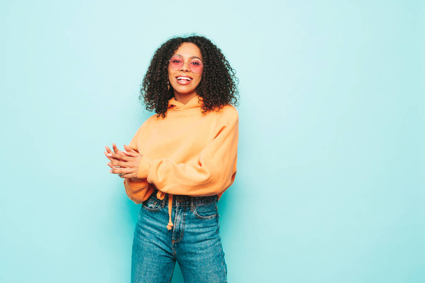 Beautiful black woman with afro curls hairstyle.Smiling model in orange hoodie and trendy jeans clothes. Sexy carefree female posing near blue wall in studio in sunglasses - Foto, Imagem