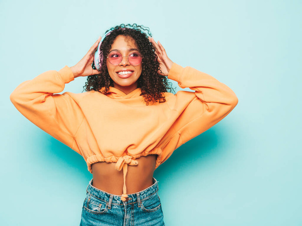 Beautiful black woman with afro curls hairstyle.Smiling model in orange hoodie and jeans.Sexy carefree female listening music in wireless headphones.Posing in studio near blue wall - Photo, image