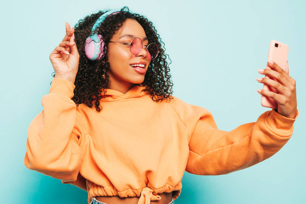 Beautiful black woman with afro curls hairstyle.Smiling model in orange hoodie and jeans.Sexy carefree female listening music in wireless headphones.Posing in studio near blue wall.Taking selfie - Foto, immagini