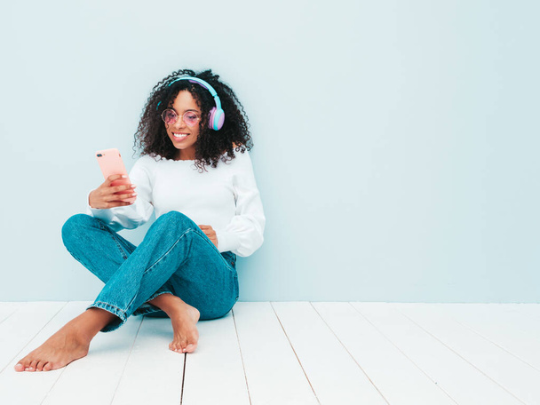 Beautiful black woman with afro curls hairstyle.Smiling model in sweater and jeans.Sexy carefree female listening music in wireless headphones.Sitting in studio near light blue wall.Taking selfie - Foto, immagini