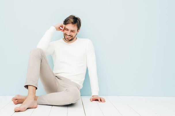 Portrait of handsome smiling hipster lumbersexual businessman model wearing casual white sweater and trousers. Fashion stylish man posing against light blue wall in studio - Photo, Image