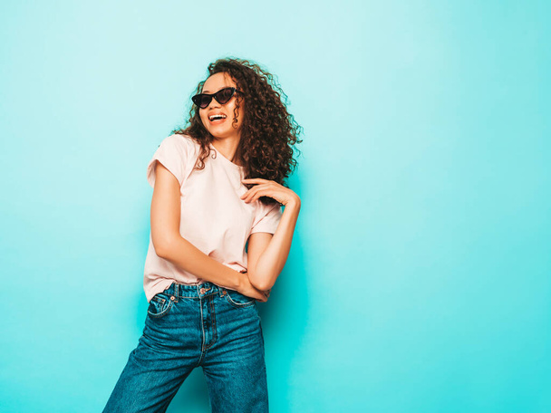 Beautiful black woman with afro curls hairstyle.Smiling model in white trendy t-shirt and jeans clothes. Sexy carefree female posing near blue in studio in sunglasses. Tanned and cheerful  - Φωτογραφία, εικόνα