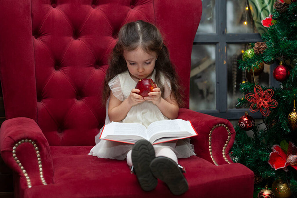 Little girl reads a book in a chair near the Christmas tree. The child eats an apple. - Φωτογραφία, εικόνα