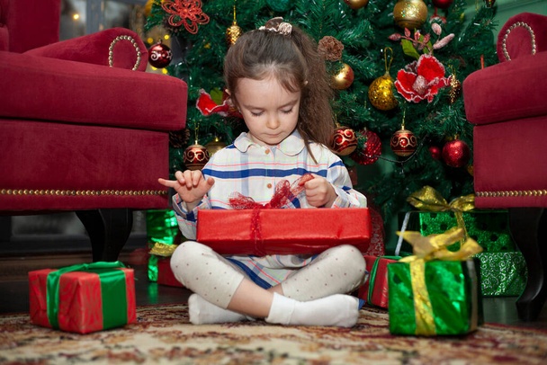 A little girl opens gifts near the Christmas tree. New Year's morning. Child and New Year's gifts - Photo, Image