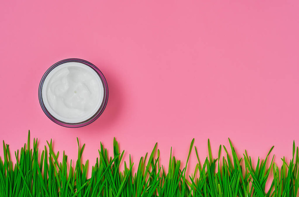 Crème cosmétique dans un bocal, fond rose vif avec herbe verte. Pose plate, vue de dessus avec espace de copie. Appliquer une crème hydratante bio. Concept de soins des mains ou du visage - Photo, image