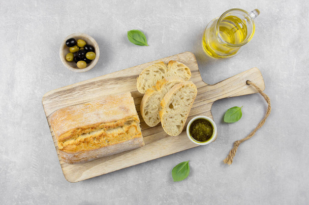 Tuore italialainen ciabatta-leipä, jossa yrttejä, oliiviöljyä, mustia ja vihreitä oliiveja, basilikanlehtiä ja pestokastiketta kevyellä betonitaustalla. Yläkuva.  - Valokuva, kuva