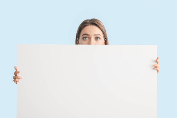 Surprised young woman with blank poster on color background - Photo, Image