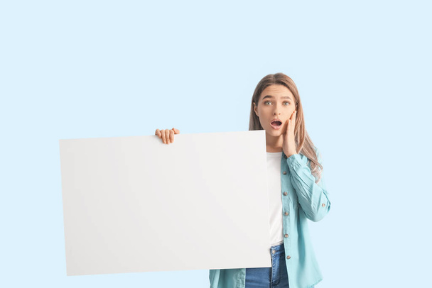 Surprised young woman with blank poster on color background - Fotografie, Obrázek