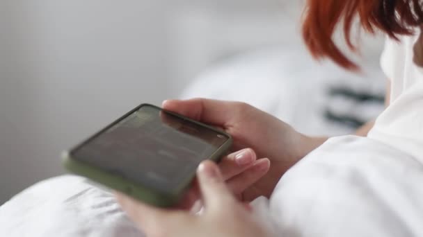 Vrouwelijke hand houden smartphone en kijken naar video zitten op een bed binnen huis - Video