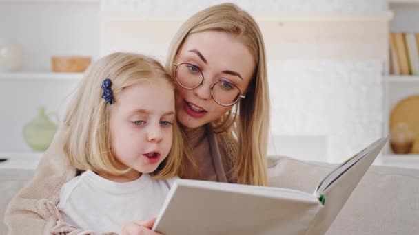 Portrait of two people family young single mother blonde woman mum wearing glasses caring mom reads literary book fairy tale to little daughter child toddler preschool kid sitting at home on weekend - Footage, Video