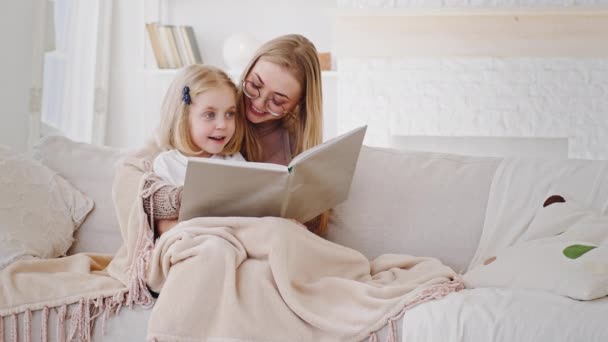 Smart adult mother babysitter reading book literature fairy tale to little daughter schoolgirl preschool child kid sitting together covered with blanket at home on couch discussing talking laughing - Footage, Video