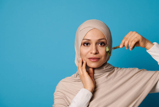 Beautiful Muslim woman in hijab massaging her face with jade roller. Isolated beauty portrait on blue background with space for text - Foto, Bild