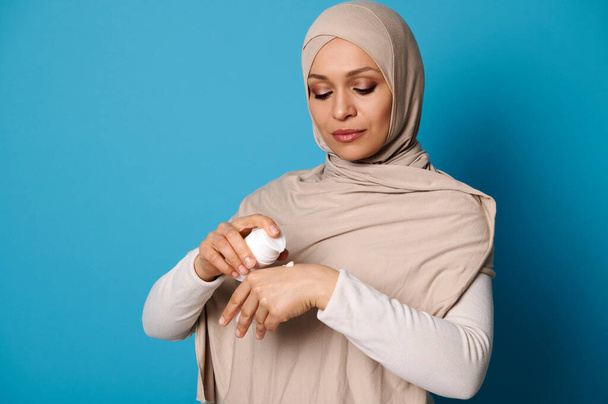Portrait of beautiful Arab Muslim woman in hijab squeezing cream on hand, isolated on blue background with space for text - Photo, image