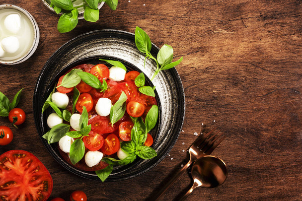 Summer italian salad Caprese with red tomatoes and mozzarella cheese with green basil leaves and olive oil dressing. Top view, wooden table - 写真・画像