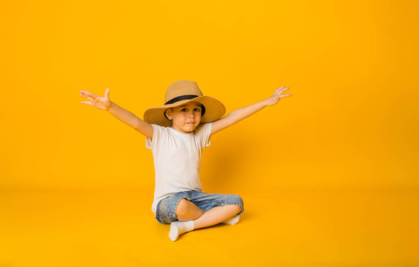 niño feliz en un sombrero de paja se sienta sobre un fondo amarillo y señala con las manos hacia un lado - Foto, imagen