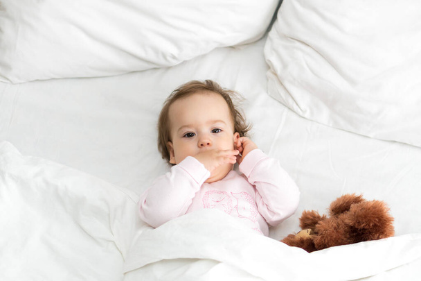 Authentic portrait cute caucasian little infant chubby baby girl or boy in pink sleepy upon waking with teddy bear looking at camera in white bed. Child care, Childhood, Parenthood, lifestyle concept - Fotoğraf, Görsel