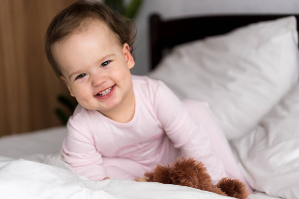 Authentic portrait caucasian little infant chubby baby girl or boy in pink sleepy upon waking looking at camera smiling cute and grimaces in white bed. Child care, Childhood, Parenthood, life concept - Foto, immagini