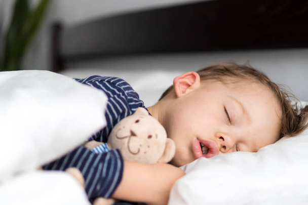 Authentic portrait sick cute caucasian little preschool baby boy in blue sleep with teddy bear on white bed. child resting at lunchtime. care, medicine and health, Childhood, Parenthood, life concept - Foto, Bild