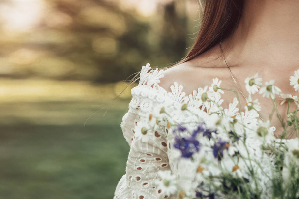 Eenzame vrouw staat alleen in de zomer stadspark of bos bossen, houden wilde bloemen. Lady gekleed in witte jurk toont haar schouder en blote sleutelbeen. Romantisch concept, wazige achtergrond. - Foto, afbeelding