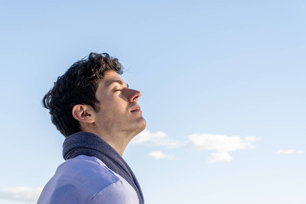Portret van een jonge jongen met hoofd naar de hemel en ogen dicht op een heldere dag met blauwe lucht en een witte wolk - Foto, afbeelding