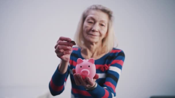 Elderly retired woman inserting coins into the piggy bank. Selective focus - Footage, Video