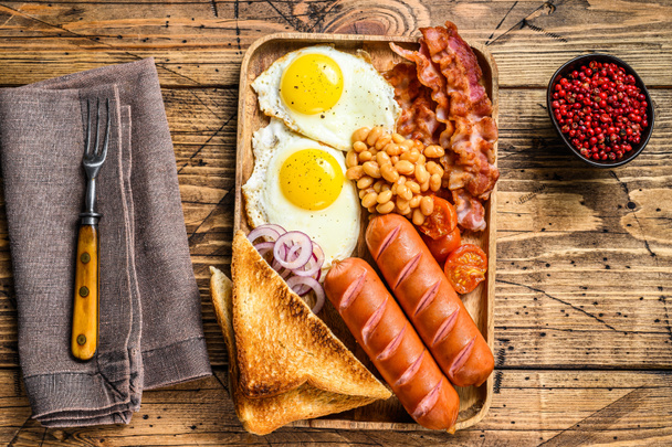 Английский завтрак в деревянной тарелке с жареными яйцами, сардельками, беконом, фасолью и тостами. деревянный фон. вид сверху - Фото, изображение