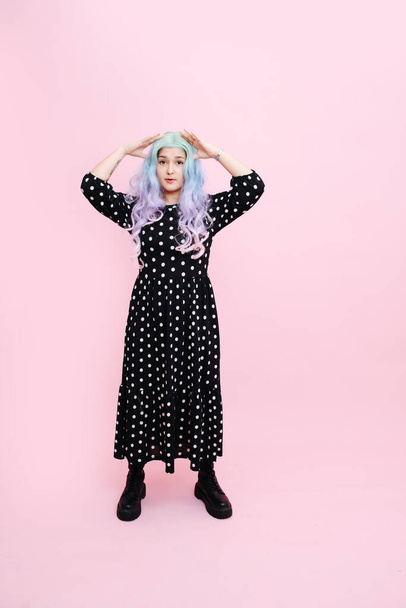 Small woman in a blue colorful wig, polka dot dress and heavy boots over pink background. She's pressing thumbs against her temples. - Foto, Bild