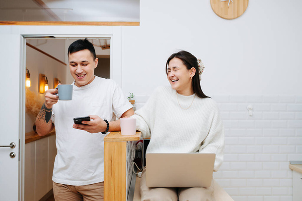 Couple joking in the kitchen. They are holding their devices and talking. Dressed casually in white and beige. She's sitting on a table, he's standing right next to her. - Φωτογραφία, εικόνα
