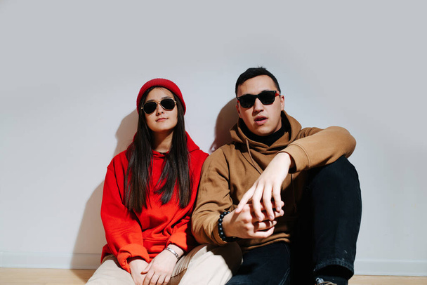 Fashionable cool-looking young couple in sunglasses and hoodies sitting on the floor shoulder to shoulder. In front of the white wall. Posing, trying to look cool. - Foto, Bild