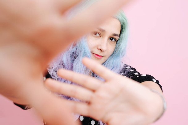 Shy woman in a blue colorful wig and polka dot dress over pink background. She's blocking view on everything except her face. - Foto, immagini