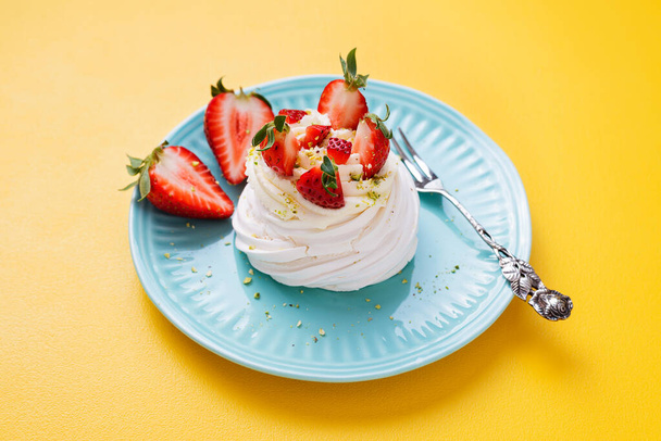 Τέλειο μίνι κέικ Pavlova με σαντιγί μασκαρπόνε και φρέσκες φέτες φράουλας, πασπαλισμένες με θρυμματισμένα φιστίκια. Κίτρινο τραπέζι - Φωτογραφία, εικόνα
