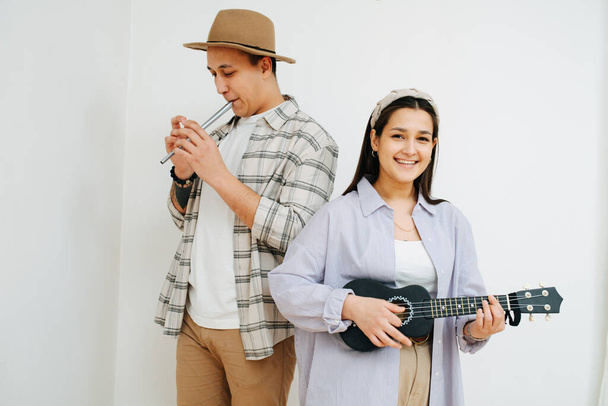 Cheerful man and woman playing thier instruments inside a room. In front of the white wall. He plays irish whistle flute, she plays small 4 string guitar. - Foto, Imagen