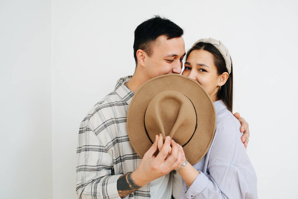 Shy young couple standing close to each other, hiding their lips behind a hat. In front of the white wall. Man hugs woman. - Photo, image