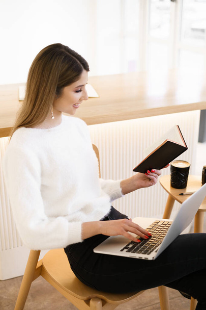 Mujer joven en la cafetería usando el ordenador portátil y mientras busca en una agenda negra - Foto, Imagen