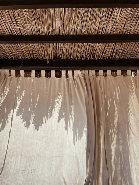 Όμορφο σαλόνι περίπτερο θόλο για να χαλαρώσετε στην παραλία. Πολυτελείς καλοκαιρινές διακοπές σε τροπικό θέρετρο παράδεισο ξενοδοχείο. Palm φύλλα ήλιο σκιά θολή αντανάκλαση σιλουέτα σε ουδέτερο μπεζ κουρτίνες - Φωτογραφία, εικόνα