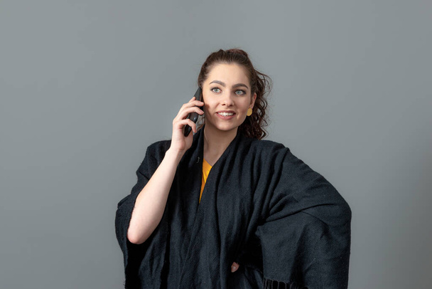 kiharatukkainen emotionaalinen nuori nainen mustassa viitta puhua matkapuhelimeen, eristetty harmaalla taustalla - Valokuva, kuva
