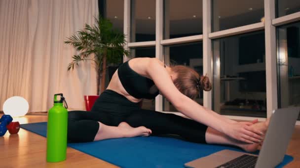 Mujer joven estirándose mientras hace yoga. Mirando el portátil. Entrenamiento interior en apartamento, cuarentena. Noche. - Imágenes, Vídeo