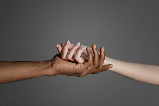 White caucasian hand in black afro american hand holding together on grey background. - Zdjęcie, obraz