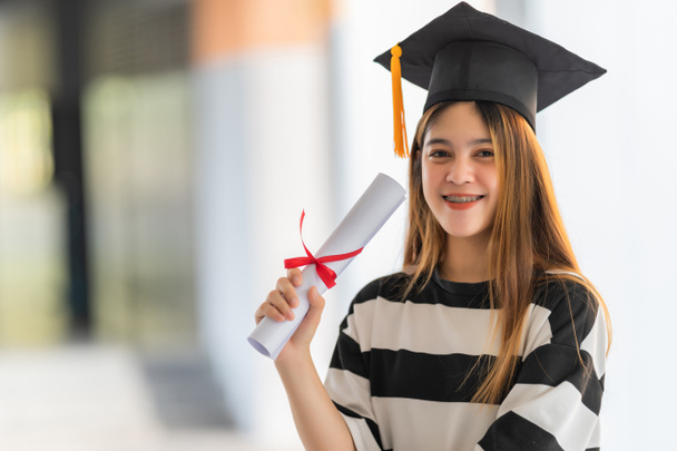 Młoda azjatycka kobieta absolwentka uniwersytetu w sukni dyplomowej i moździerz posiada certyfikat ukończenia studiów świętuje osiągnięcia edukacyjne w kampusie uniwersyteckim. Zdjęcie edukacyjne - Zdjęcie, obraz