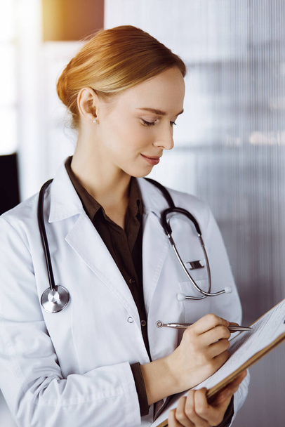 Cheerful smiling female doctor using clipboard in clinic. Portrait of friendly physician woman at work. Medical service in hospital. Medicine concept - Photo, Image