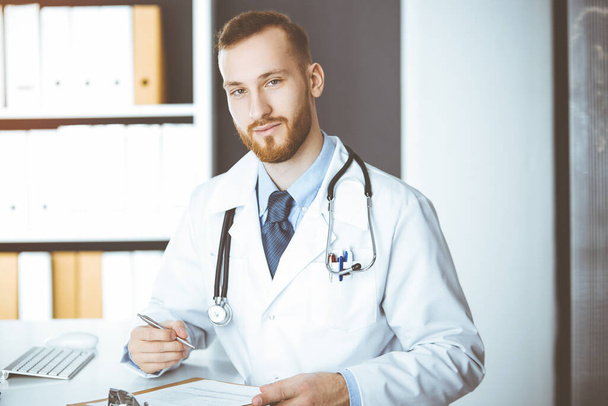 Friendly red-bearded doctor sitting and writing at clipboard in sunny clinic. Medicine concept, pandemic stop - Foto, Imagen