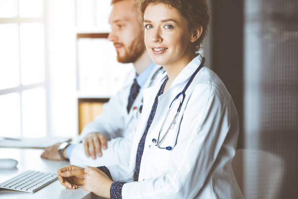 Friendly female doctor sitting in clinic with her male colleague at the background in sunny clinic. Medicine concept - 写真・画像