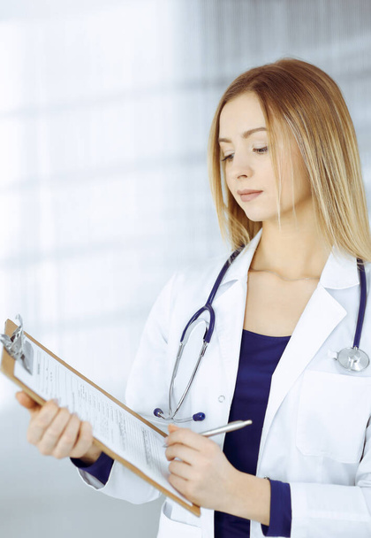 Young intelligent woman-doctor is prescribing some medicine for a patient, using a clipboard, while standing in the cabinet in a clinic. Female physician with a stethoscope at her working place - Foto, Bild