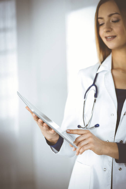 Young woman-doctor is holding a tablet computer in her hands, while standing in a clinic. Portrait of friendly female physician with a stethoscope at work. Perfect medical service in a hospital - Foto, Bild