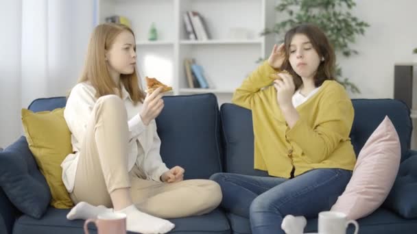 Two female friends chatting on couch and eating pizza, enjoying time together - Video, Çekim